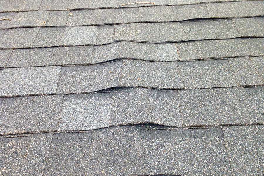 An educational slider graphic titled The Old 15-Year Life Cycle showcasing a close-up section of a gray shingle roof where multiple rows are visibly warping, buckling, and lifting due to natural material exhaustion.