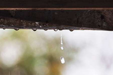 An educational slider module image illustrating drainage failure, showcasing rainwater actively dripping and spilling over the edge of a dark residential gutter line.
