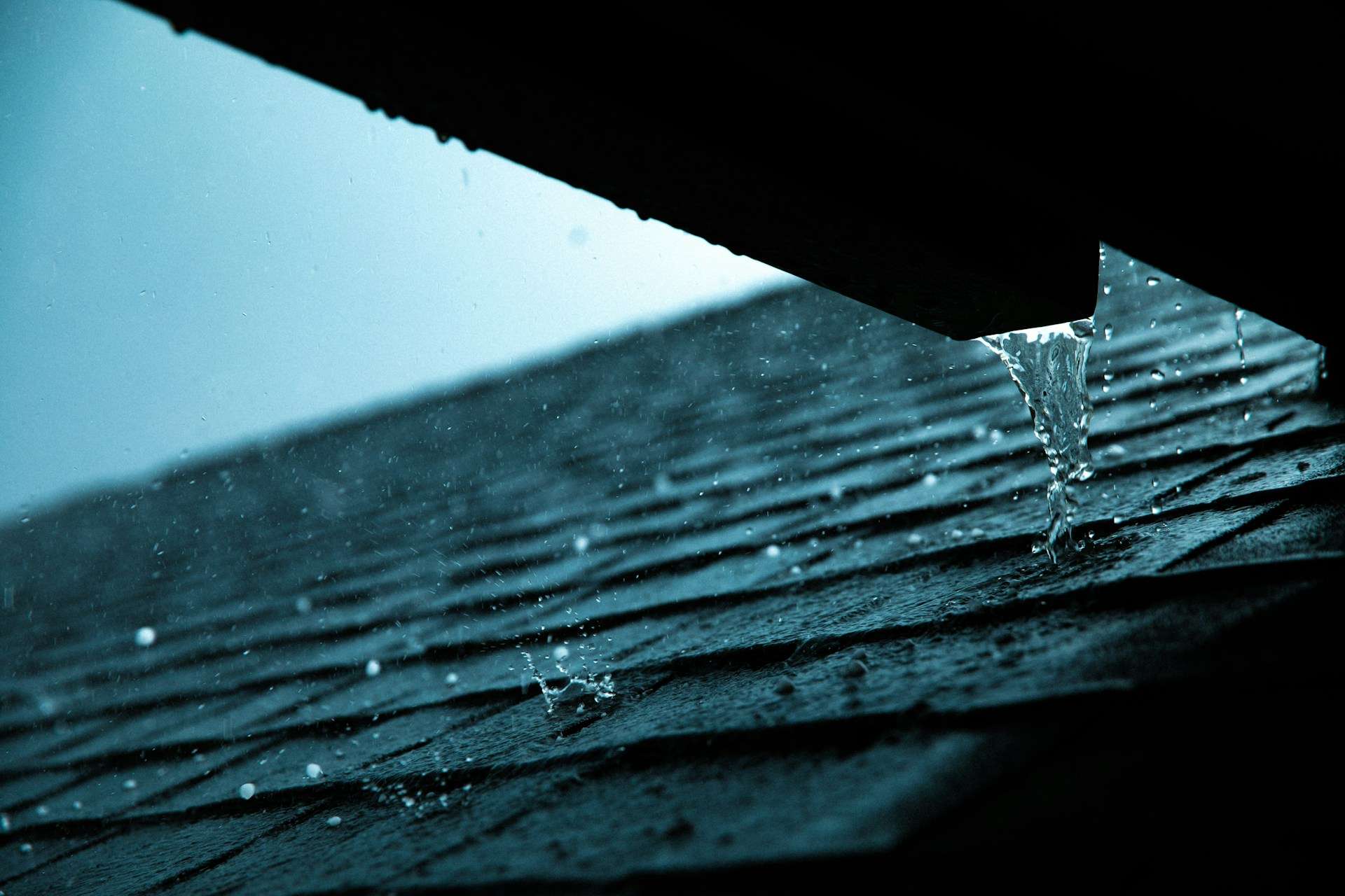 An educational module image showing a section of an asphalt shingle roofline where water overflows past the gutter trim due to capacity blockages or incorrect structural pitch.