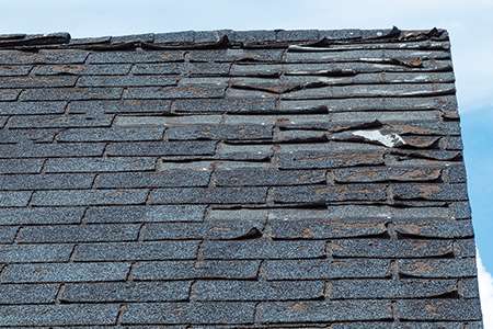 An educational slider graphic titled Physical Structural Warning Signs showing a close-up section of a weathered gray roof with severely curling, warped, and damaged architectural asphalt shingles due to advanced material fatigue.