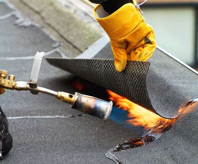 A commercial roofing technician using a propane torch tool to heat-weld the seam of a dark flat roofing membrane layer.