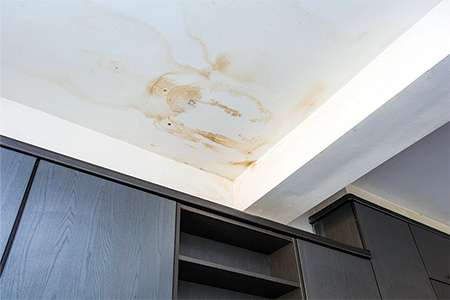 An educational slider graphic titled Active Moisture Paths displaying a prominent yellowish-brown water ring and moisture stain spreading across a white interior drywall ceiling above dark cabinets.