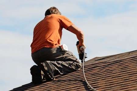 An educational slider graphic titled 1 Year Mark showing a roofer in an orange shirt kneeling on a steep architectural shingle roof facet while operating a pneumatic tool to secure materials.