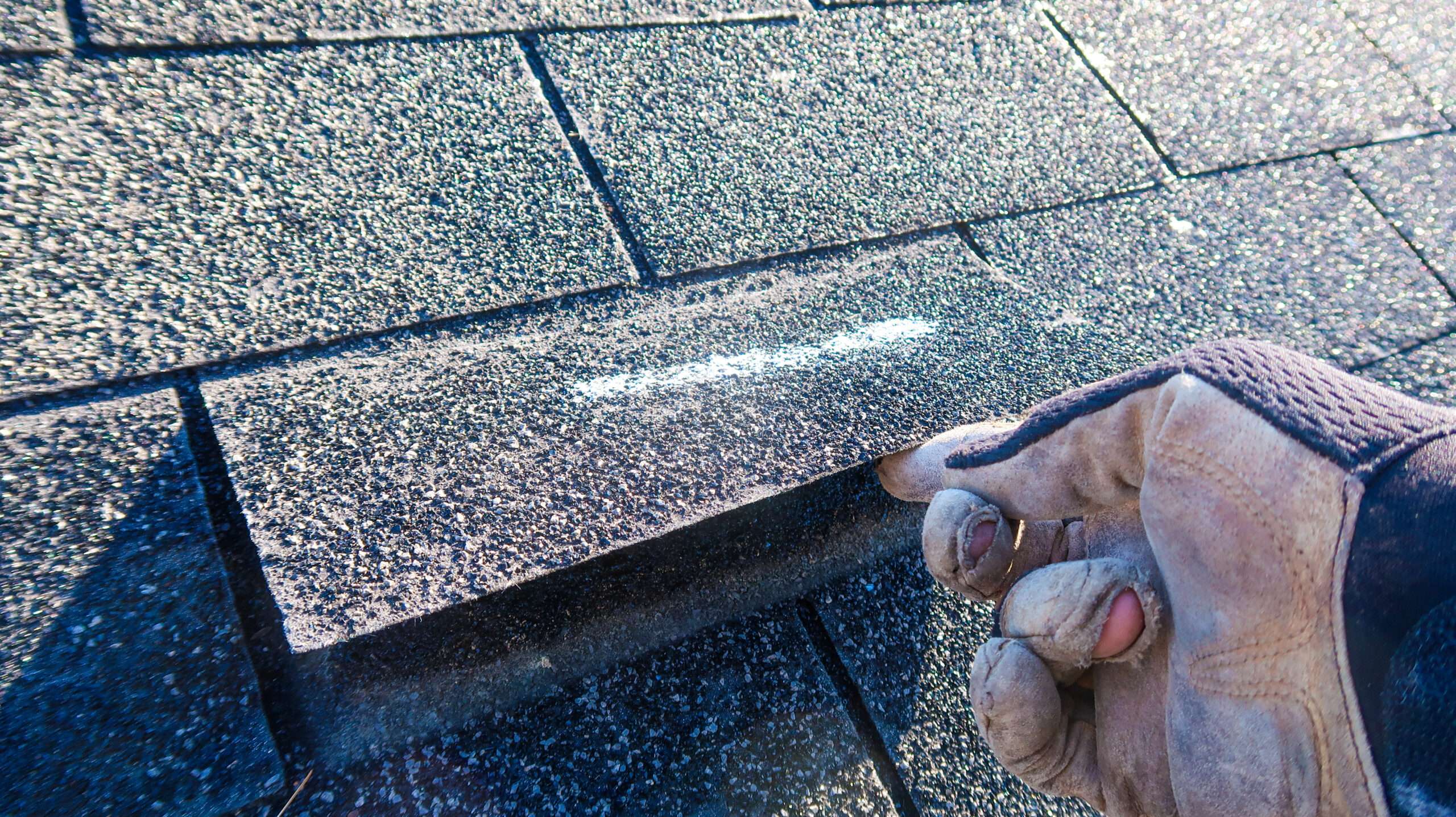 A gloved hand lifting an asphalt shingle to check for wind uplift damage during a roof inspection.