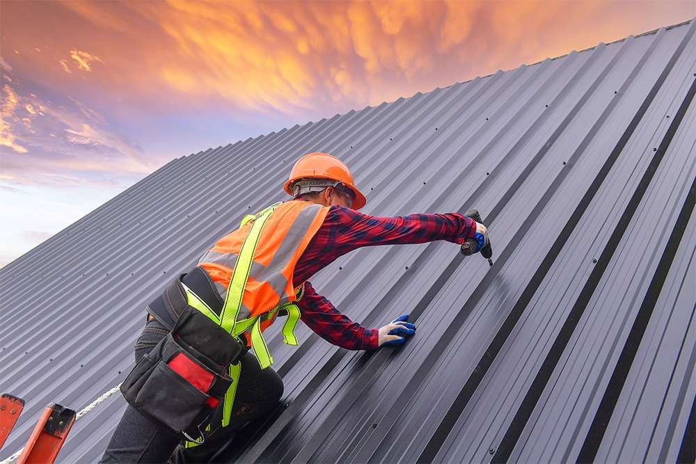 A construction worker wearing a safety harness and hard hat secures grey corrugated metal roofing panels on a steep commercial roof slope.